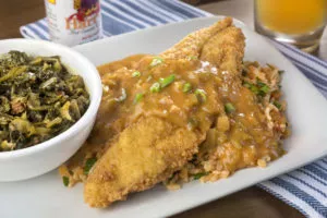 A fried fish platter with two crispy fillets smothered in gravy and green onions, served with rice pilaf and a side of collard greens. Harry's Hot Sauce is in the background.