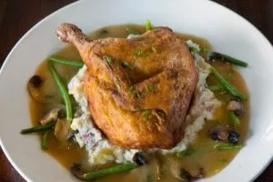 A roasted chicken leg quarter with crispy skin, served over mashed potatoes, surrounded by a rich brown gravy with green beans and sliced mushrooms, on a white plate.