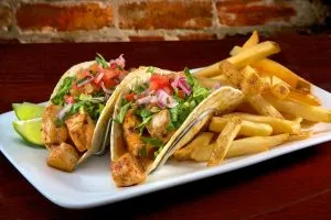 A plate with two chicken tacos, topped with lettuce, salsa, and lime wedges, served alongside a pile of golden french fries, all against a blurred brick background.