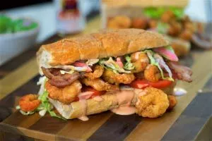 A Po' Boy sandwich on a wooden board, filled with crispy fried shrimp, bacon, tomato, lettuce, pickled red onions, and a creamy orange sauce.