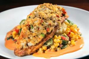A pan-seared fish fillet topped with crabmeat stuffing and a succotash of corn, okra, and bell peppers, served with a creamy orange sauce on a white plate.